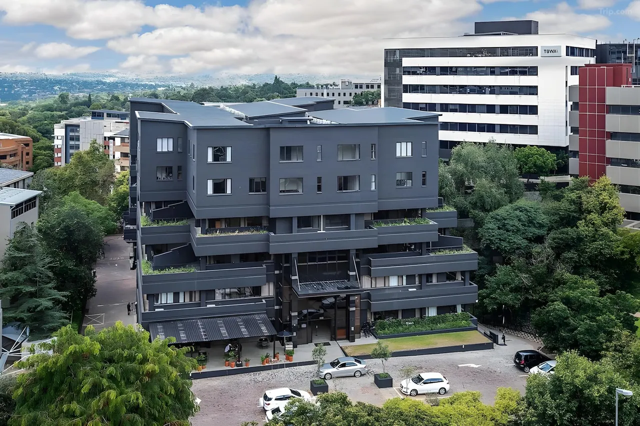 Aerial view of a modern charcoal-brick apartment building with stepped, planted balconies, surrounded by trees with the leafy Sandton skyline behind