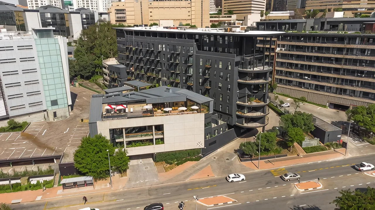 Aerial view of a modern dark-clad apartment block with private balconies, a rooftop terrace with umbrellas and a curved glass stairwell, set among Sandton's high-rise skyline